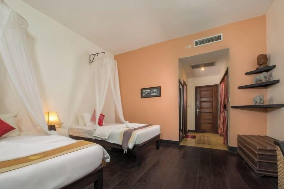 Een hotelkamer met twee bedden, een nachtkastje met een lamp, een foto aan de muur, een deur naar een andere kamer en een plank met decoratieve objecten.
