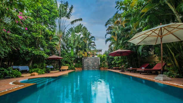 A swimming pool surrounded by lush greenery and palm trees, with red umbrellas and lounge chairs. - Marirath Boutique Hotel  (Siem Reap)