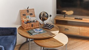 A small, round wooden table with a globe, a basket of snacks, and a magazine. A blue chair is to the left, and a wooden cabinet with a TV is in the background. - Central Cosy Harrogate Apartment (North Yorkshire)
