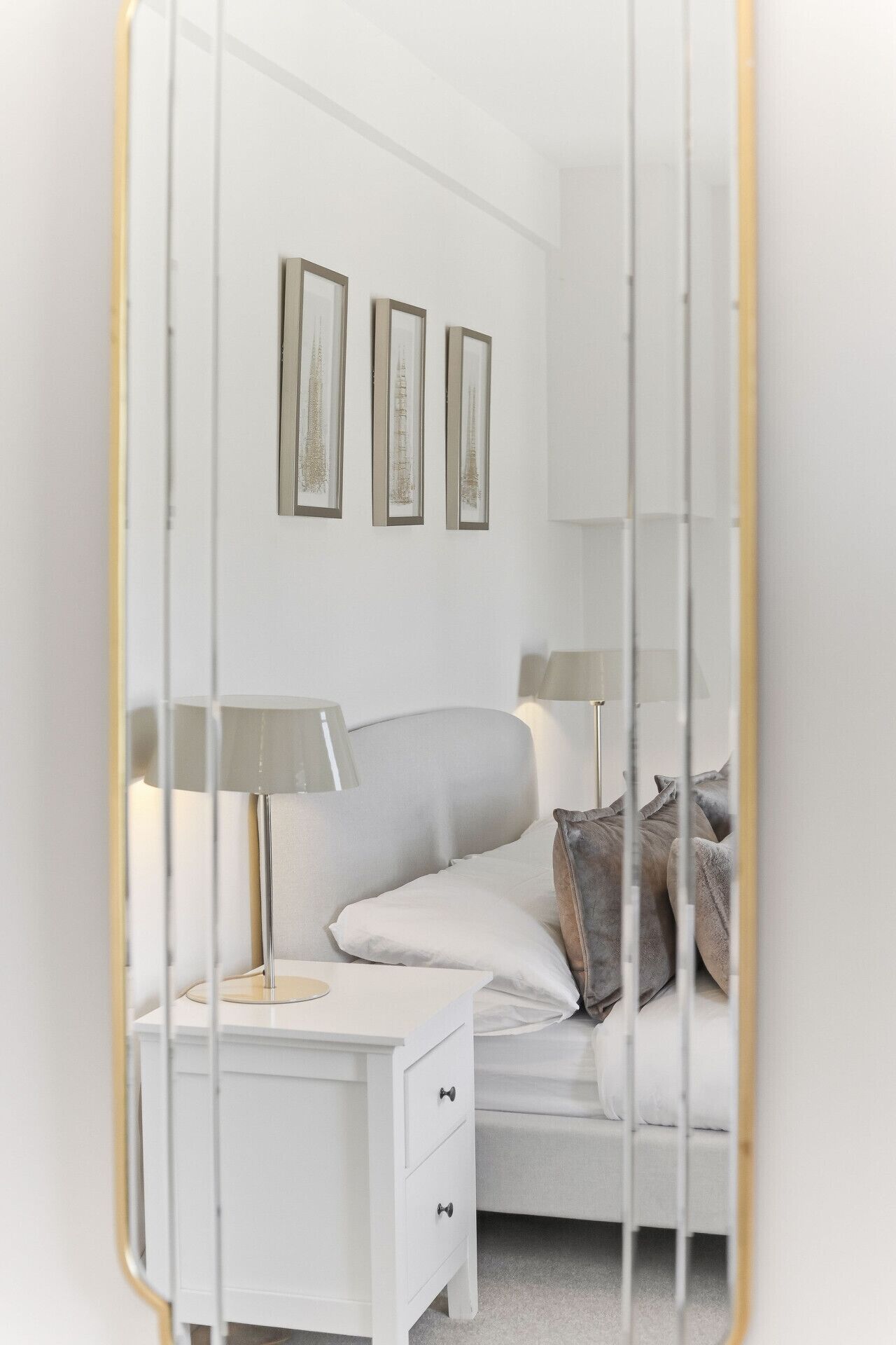 Um quarto com uma cama branca, duas luminárias de cabeceira, uma mesa de cabeceira com gaveta e três quadros na parede.