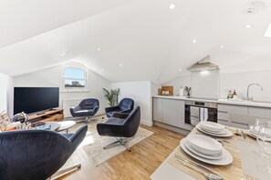 Un salon moderne doté d’un téléviseur à écran plat, de fauteuils en cuir noir, d’une table à manger dressée avec de l’assiette et des couverts, et d’une cuisine équipée d’une cuisinière et d’un four.