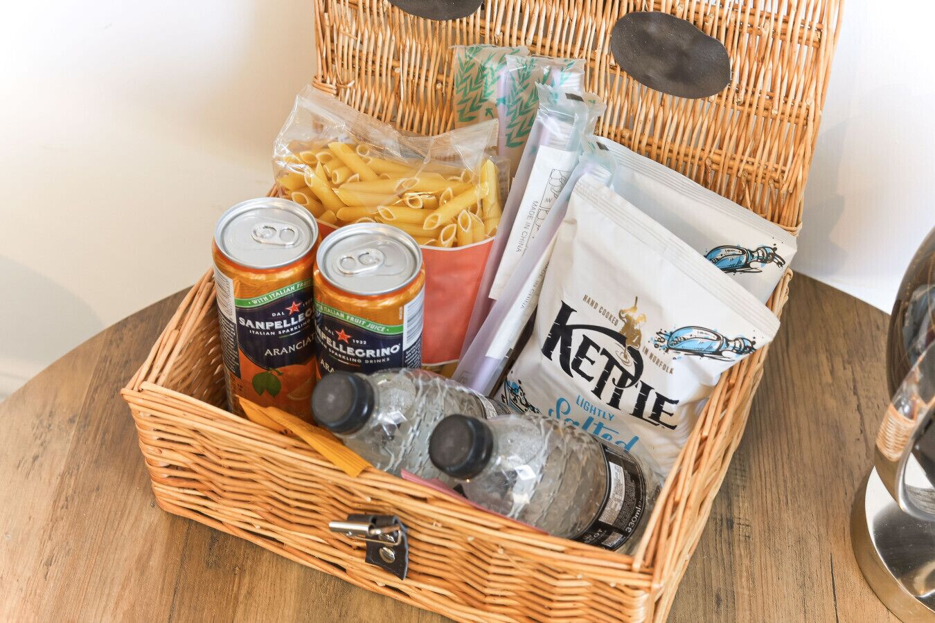 Una cesta de mimbre con pasta, bebidas enlatadas, agua embotellada y una bolsa de aperitivos.