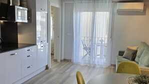 Una cocina moderna con armarios blancos y una estufa negra, un comedor con una mesa redonda y sillas amarillas, y un balcón con una cortina blanca.
