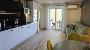 Una cocina moderna con armarios blancos, una encimera negra y un horno empotrado. Hay una zona de comedor con una mesa redonda y sillas amarillas. Se ve un sofá verde y un balcón con una silla.