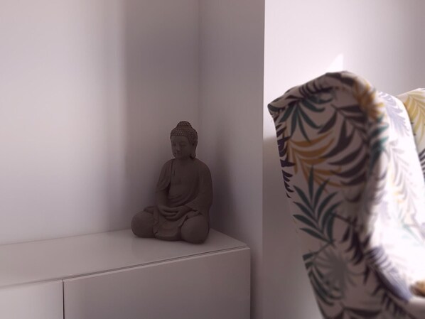 Une pièce au décor minimaliste, avec une statue de Bouddha posée sur une étagère blanche, un tissu à motifs drapé sur une chaise et un mur blanc uni.