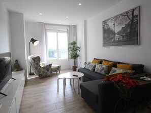 Ein modernes Wohnzimmer mit einer Couch, Sesseln, einem Couchtisch, einem Fernseher und einem großen Fenster mit Vorhängen.