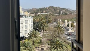 A view from a window overlooking a cityscape with palm trees, a hill with buildings, and a clear sky.