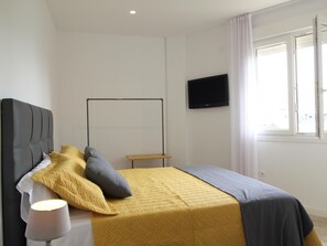 A bedroom with a bed, a bedside table, a lamp, a TV on the wall, and a window with curtains.
