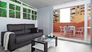 Un salon moderne avec un canapé gris, une table basse en verre et un balcon agrémenté de chaises et d’une table blanches.
