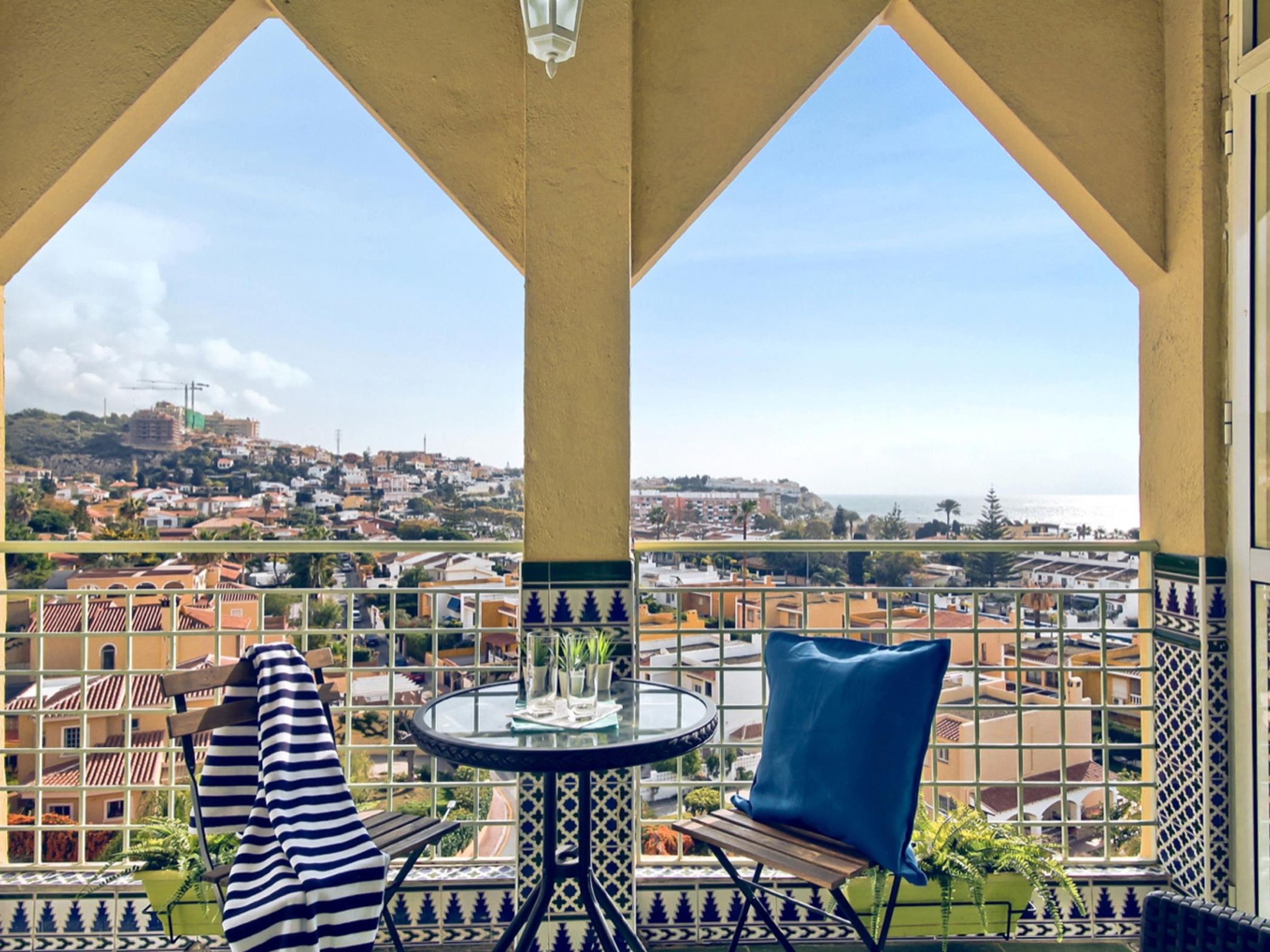 A balcony with a table and chairs, offering a view of a coastal town and the sea.