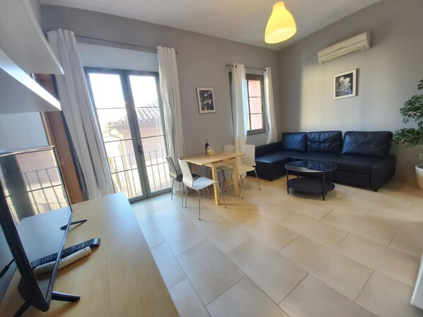 Een moderne woonkamer met een bank, een eettafel met stoelen, een klein bijzettafeltje en een groot raam met gordijnen.