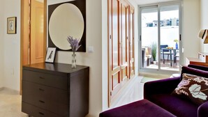 Ein modernes Wohnzimmer mit einer dunkelholznen Kommode, einem großen runden Spiegel und einem violetten Sofa mit einem floralen Kissen.