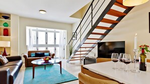 Ein modernes Wohnzimmer mit Sofa, Couchtisch und Essbereich. Eine Treppe mit Geländer führt zu einer oberen Ebene.