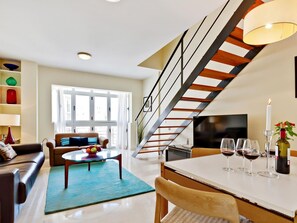 Ein modernes Wohnzimmer mit Sofa, Couchtisch und Essbereich. Eine Treppe mit Geländer führt zu einer oberen Ebene.
