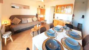 Un salon chaleureux avec un canapé, une table à manger dressée avec de l’assiette et des verres bleus, et un coin cuisine doté de meubles en bois.