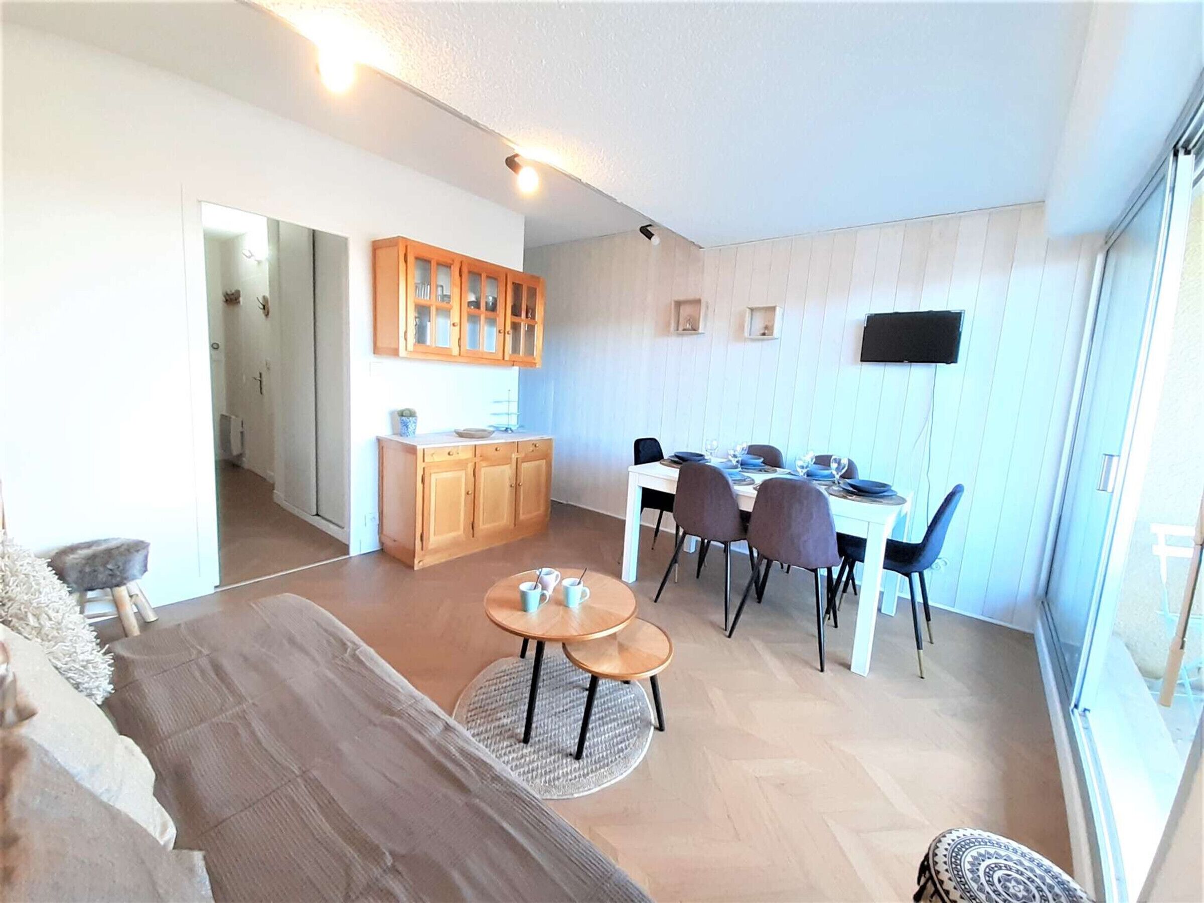 Una sala de estar moderna con un sofá, una mesa de comedor con sillas, una zona de cocina con armarios de madera y un televisor montado en la pared.
