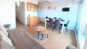 Un salon moderne comprenant un canapé, une table à manger avec des chaises, un coin cuisine avec des meubles en bois et un téléviseur fixé au mur.