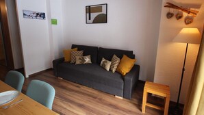 Una sala de estar con un sofá gris oscuro, dos cojines estampados, una mesita de madera, una lámpara de pie y una zona de comedor con una mesa y sillas de madera.