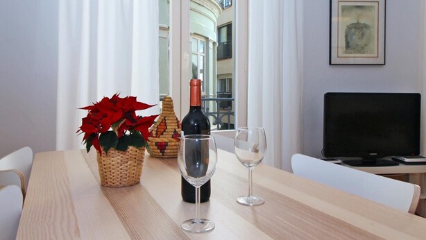 Un tavolo di legno con una bottiglia di vino, due bicchieri da vino, una pianta in vaso e un cesto di vimini. A destra, un televisore su un mobile.