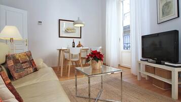 Een woonkamer met een bank, een glazen salontafel, een eettafel met stoelen, een televisie en een raam met gordijnen.