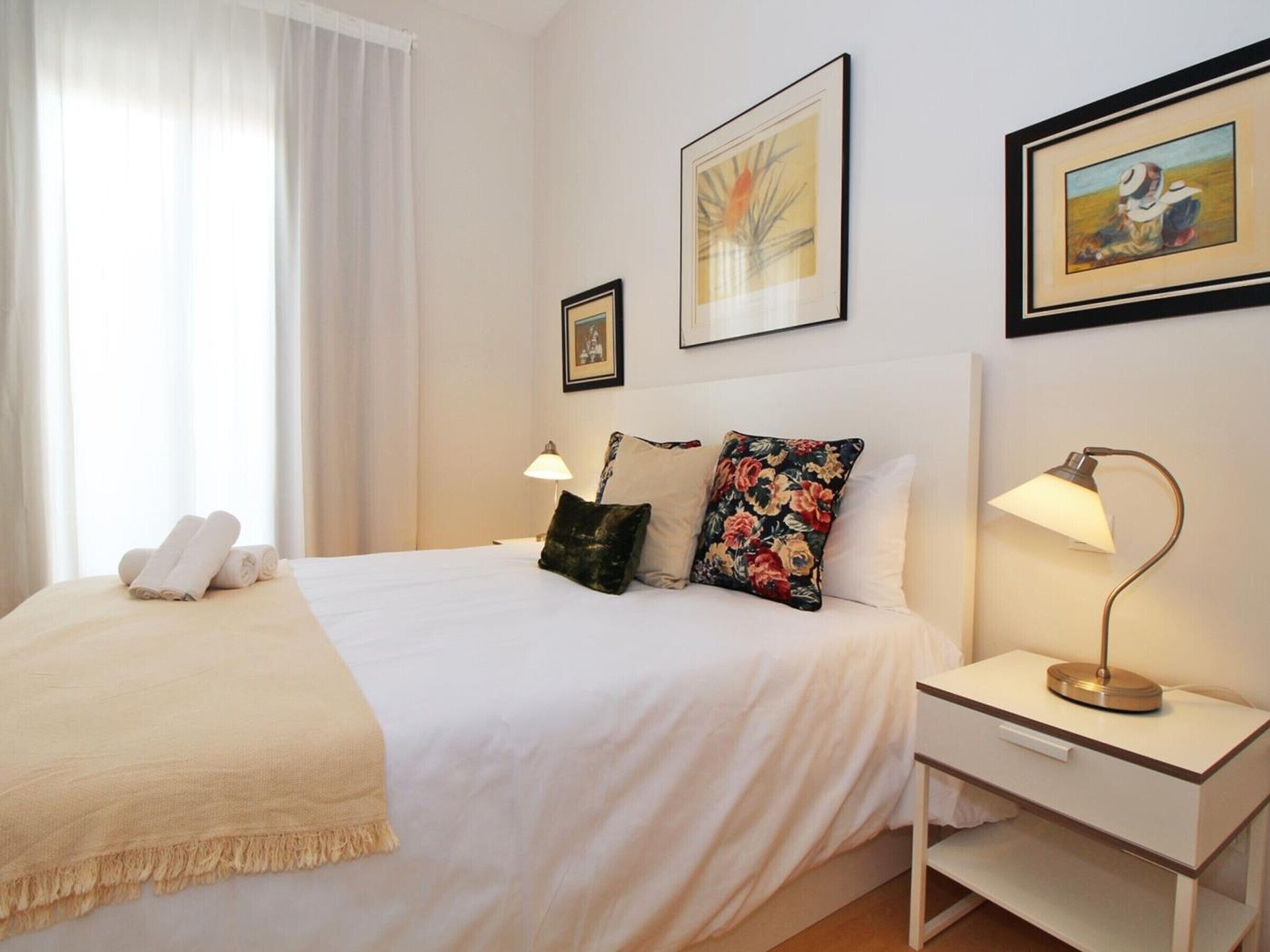 Een hotelkamer met een bed, twee bedlampen, een nachtkastje, een raam met gordijnen en twee ingelijste foto's aan de muur.
