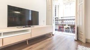 Een moderne woonkamer met een flatscreen televisie op een witte console, houten vloeren en uitzicht op een balkon met rode stoelen en een reling.