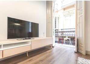A modern living room with a flat-screen TV on a white console, wooden flooring, and a view of a balcony with red chairs and a railing.