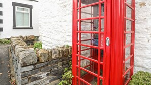 A red telephone booth in front of a white building with a stone wall and a sign for 'Cross Keys Self Catering Holiday Cottage'. - Llys Mair (Bala)
