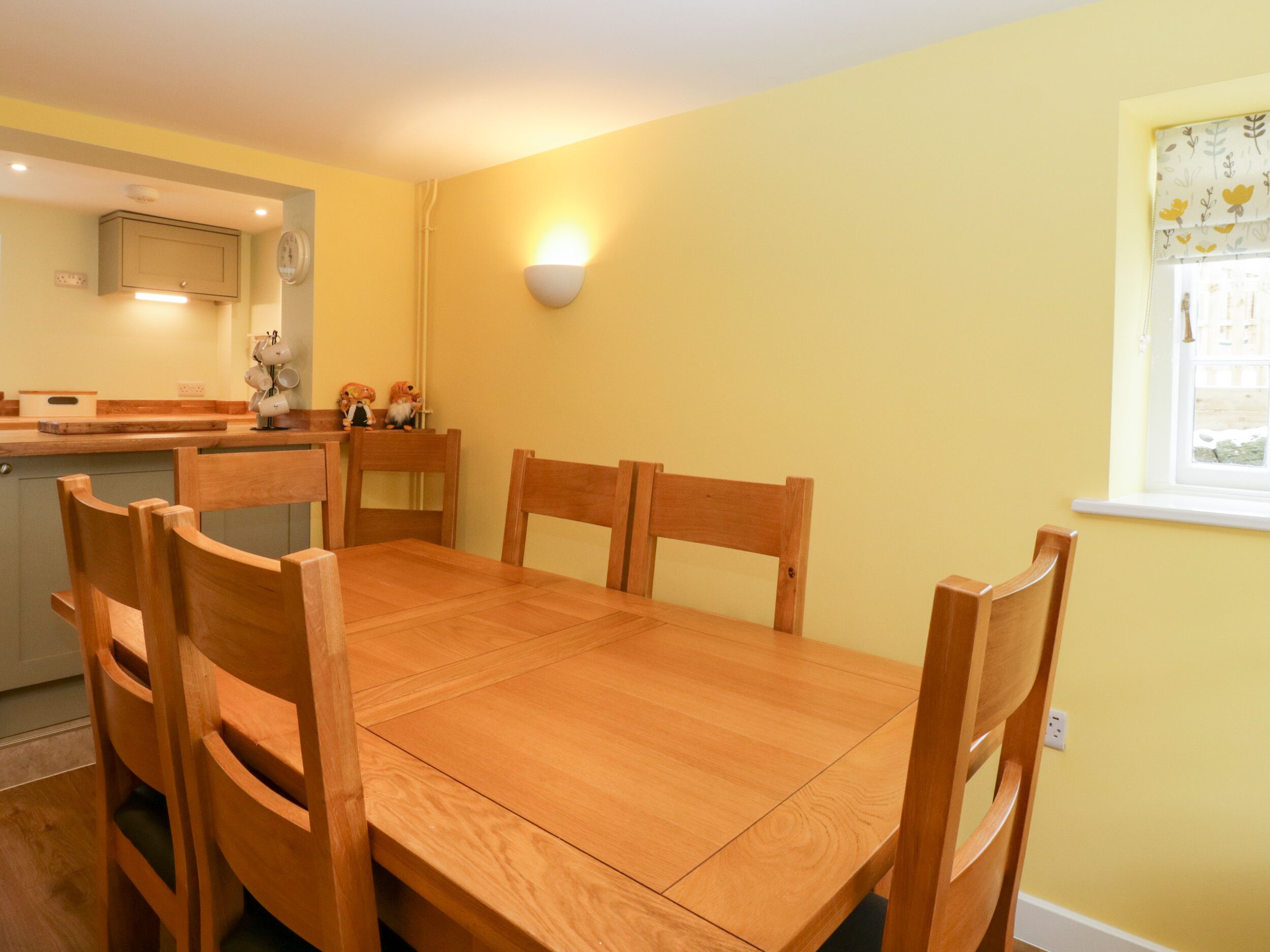 A dining area with a wooden table and chairs, a kitchen in the background, and a wall-mounted light fixture.