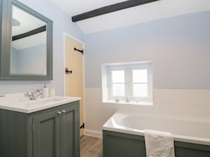 A modern bathroom with a large bathtub, a vanity with a mirror, and a window above the sink. - Granary Cottage (Warminster)