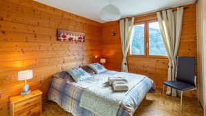Un dormitorio de cabaña de madera con una cama, una silla, una mesita de noche, una lámpara y una ventana con cortinas.