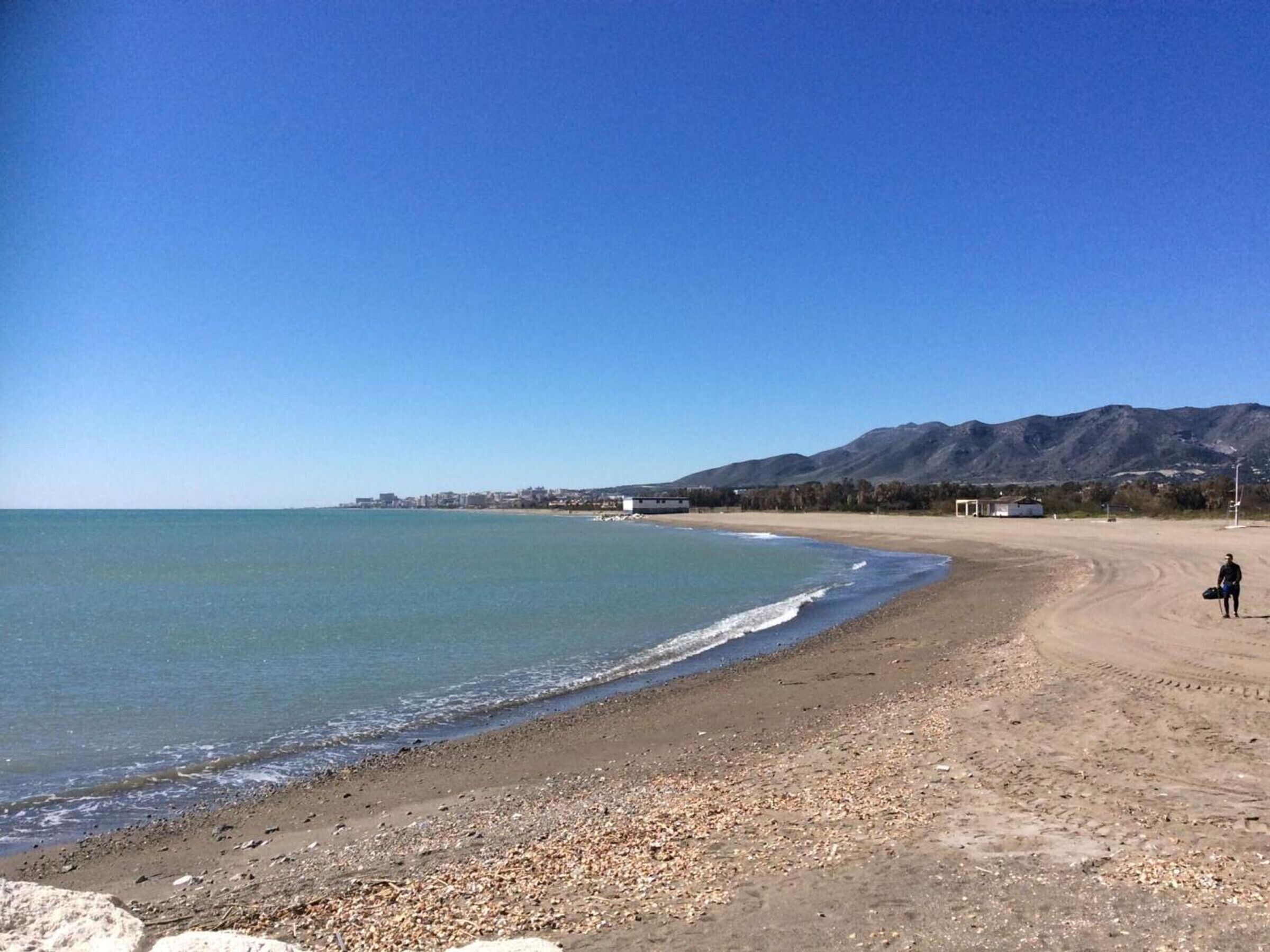 Ein Strand mit blauem Himmel, eine Person geht spazieren, und Berge im Hintergrund.