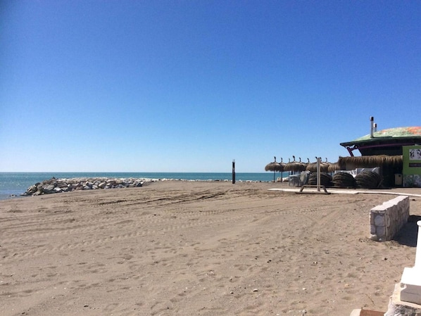 A beach with a thatched-roof structure, a sandy area, and a clear blue sky.