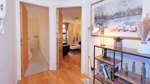 A hallway with a wooden floor, a white door, a framed painting, a lamp, and a shelf with books and decorative items. - Riverview-2br-sleeps4-citycentre-parking-fastwifi (Aberdeen)