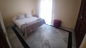 A hotel room with a bed, a nightstand, a lamp, a window with curtains, and an air conditioning unit. - Beachfront Estepona Apartment with Pool, Terrace & Free Parking (Estepona)
