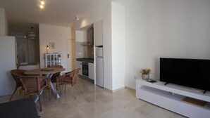 Uma sala de estar moderna com uma TV de tela plana sobre um suporte branco, área de jantar com cadeiras de vime e cozinha com armários brancos e eletrodomésticos de aço inoxidável.