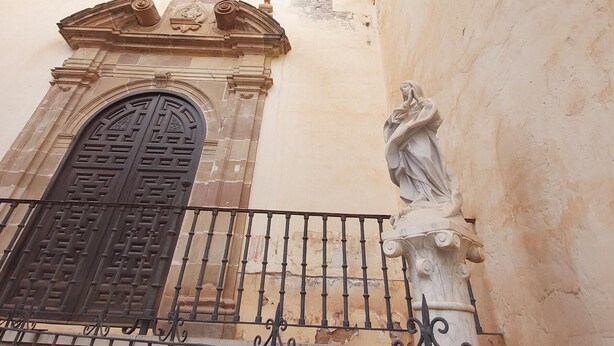 Fachada de edifício com uma grande porta em arco, uma estátua e uma varanda com grade de ferro forjado.
