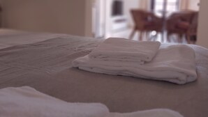 Cama bem arrumada com toalhas brancas dobradas, quarto limpo e moderno com porta visível e cadeiras ao fundo.