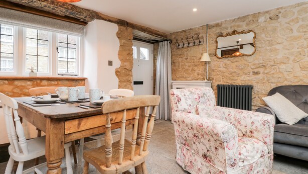 A cozy living room with a stone wall, a wooden dining table, floral armchairs, and a grey sofa. - Foxden (Sherborne)