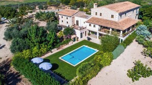 A large, two-story house with a swimming pool, surrounded by lush greenery and a sandy beach.