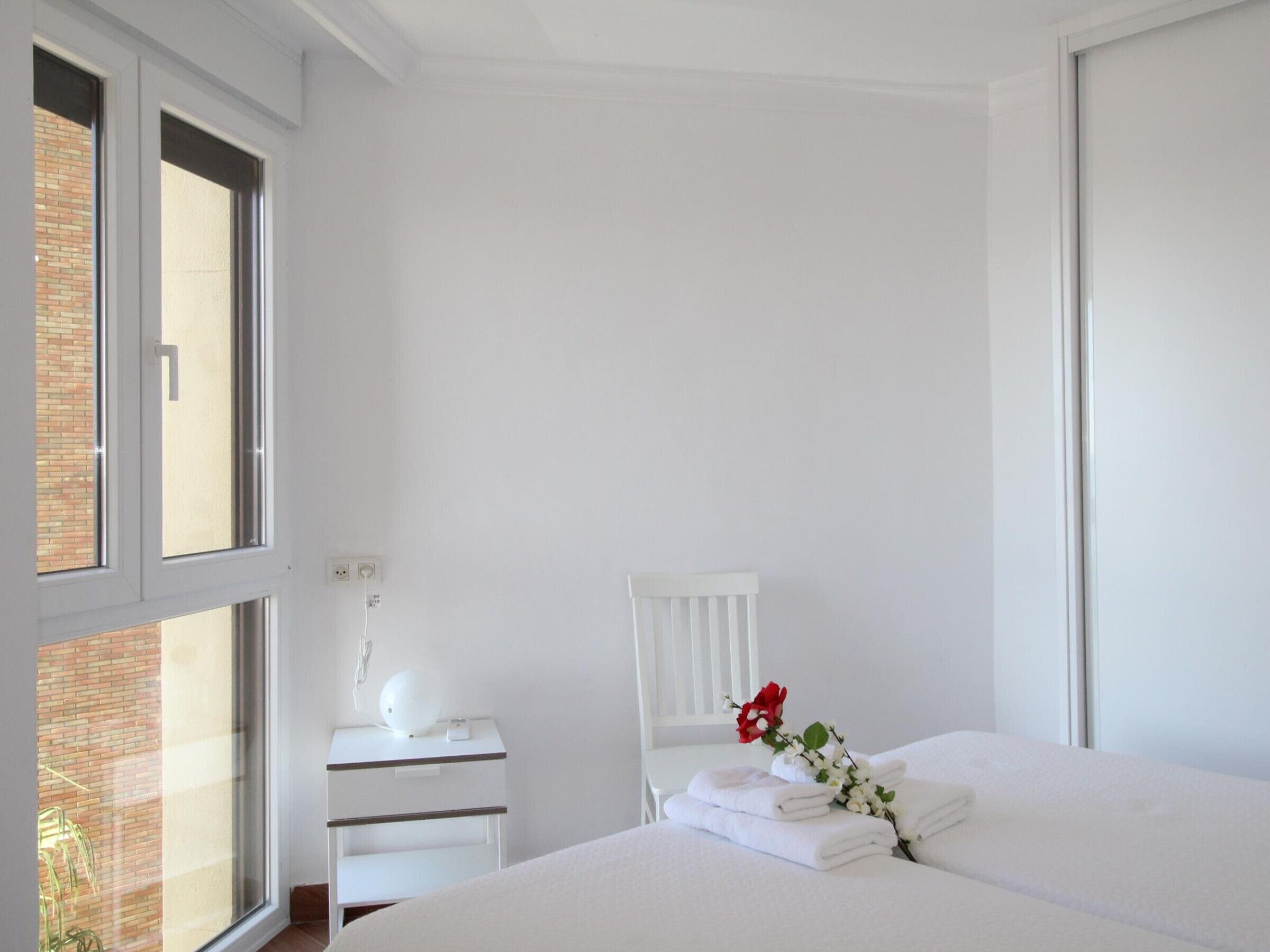 Une chambre d’hôtel avec une grande fenêtre, une table de chevet avec une lampe, une chaise et un lit avec des serviettes soigneusement pliées et une fleur rouge.