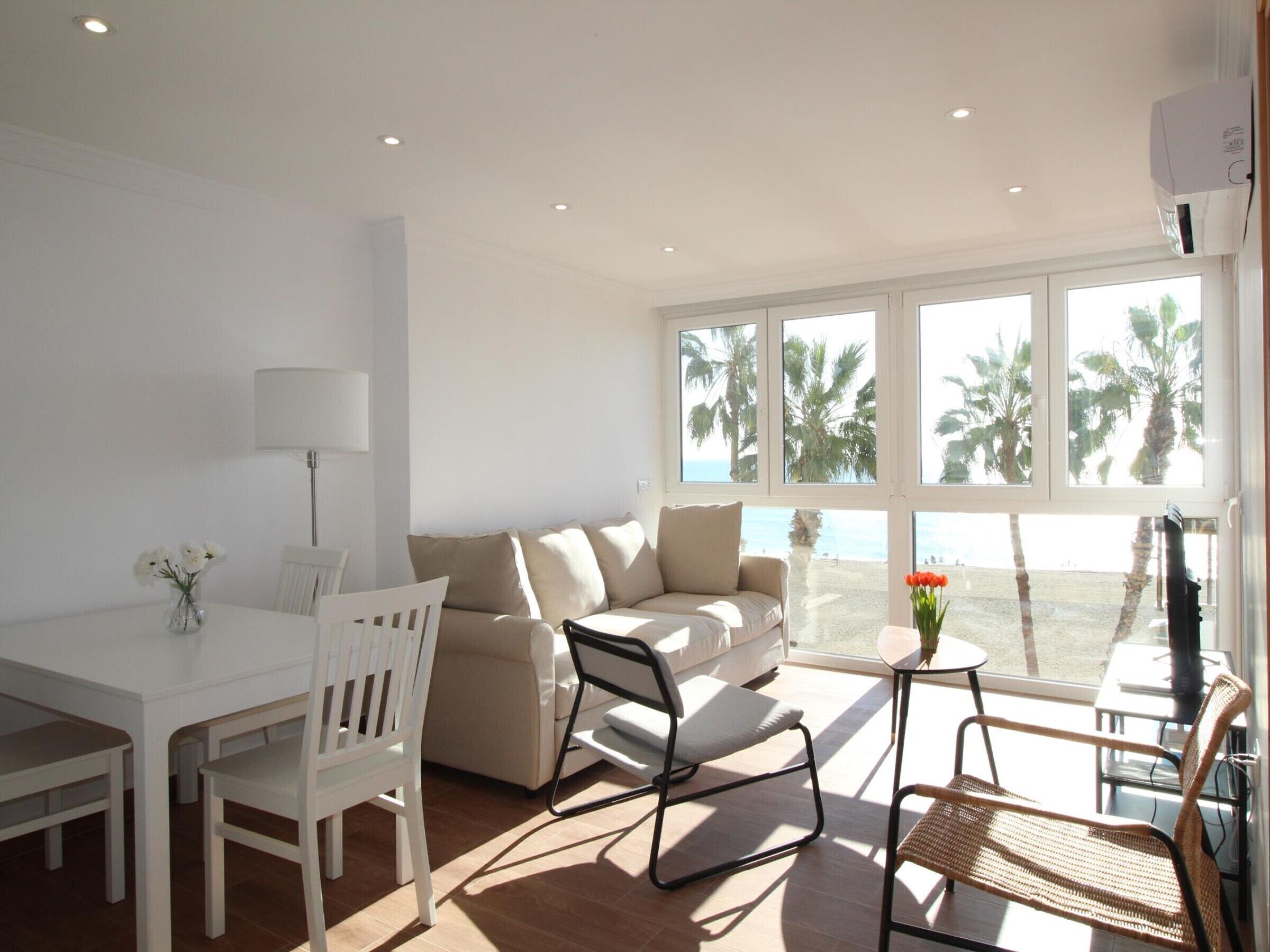 Ein heller Wohnraum mit Sofa, Sesseln, einem Tisch und Blick auf Palmen und das Meer durch große Fenster.