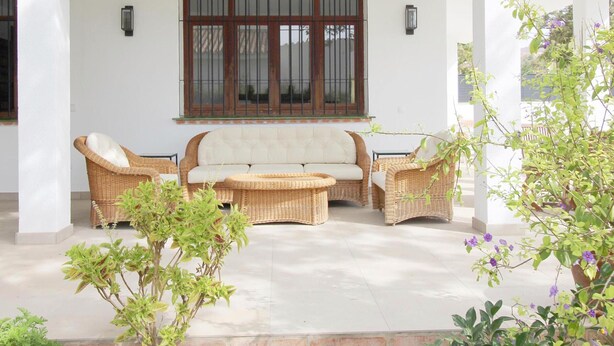 A covered patio area with a sofa and two armchairs, surrounded by potted plants and a window with bars. - Luxury Villa with Private Pool, Near Beach and Golf (Málaga)