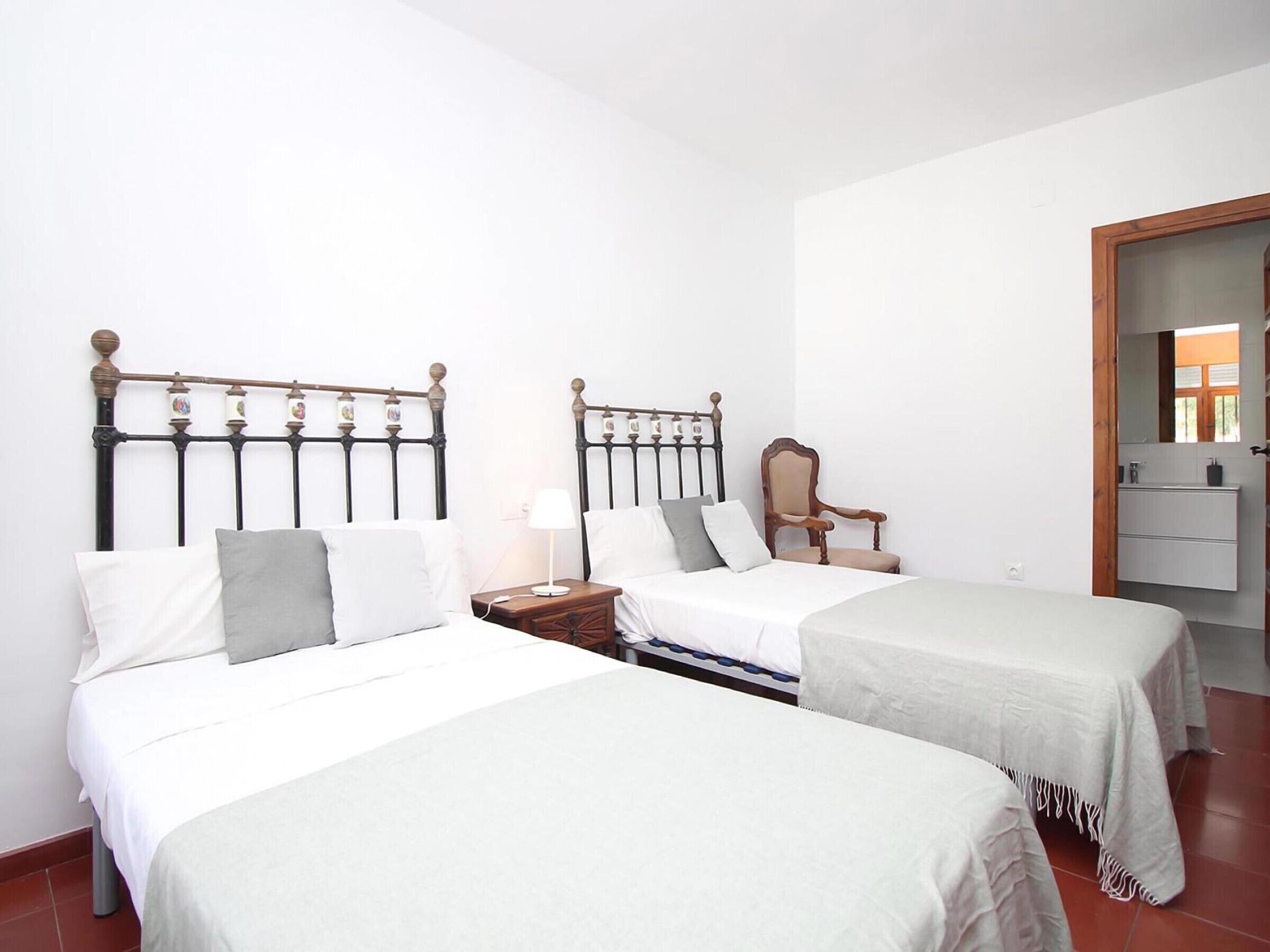 A hotel room with two beds, a wooden chair, a bedside table with a lamp, and a bathroom visible through an open door.