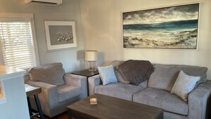 Sala de estar con un sofá gris, una mesa de centro de madera y una lámpara sobre una mesita auxiliar. Se observa un cuadro en la pared y una ventana con persianas.