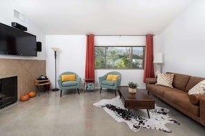 Ein modernes Wohnzimmer mit Kamin, zwei Sesseln, einer Couch, einem Couchtisch und einem großen Fenster mit Vorhängen.