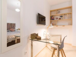 A modern bedroom with a glass desk, a chair, a bed with a colorful headboard, and a shelf with decorative items.