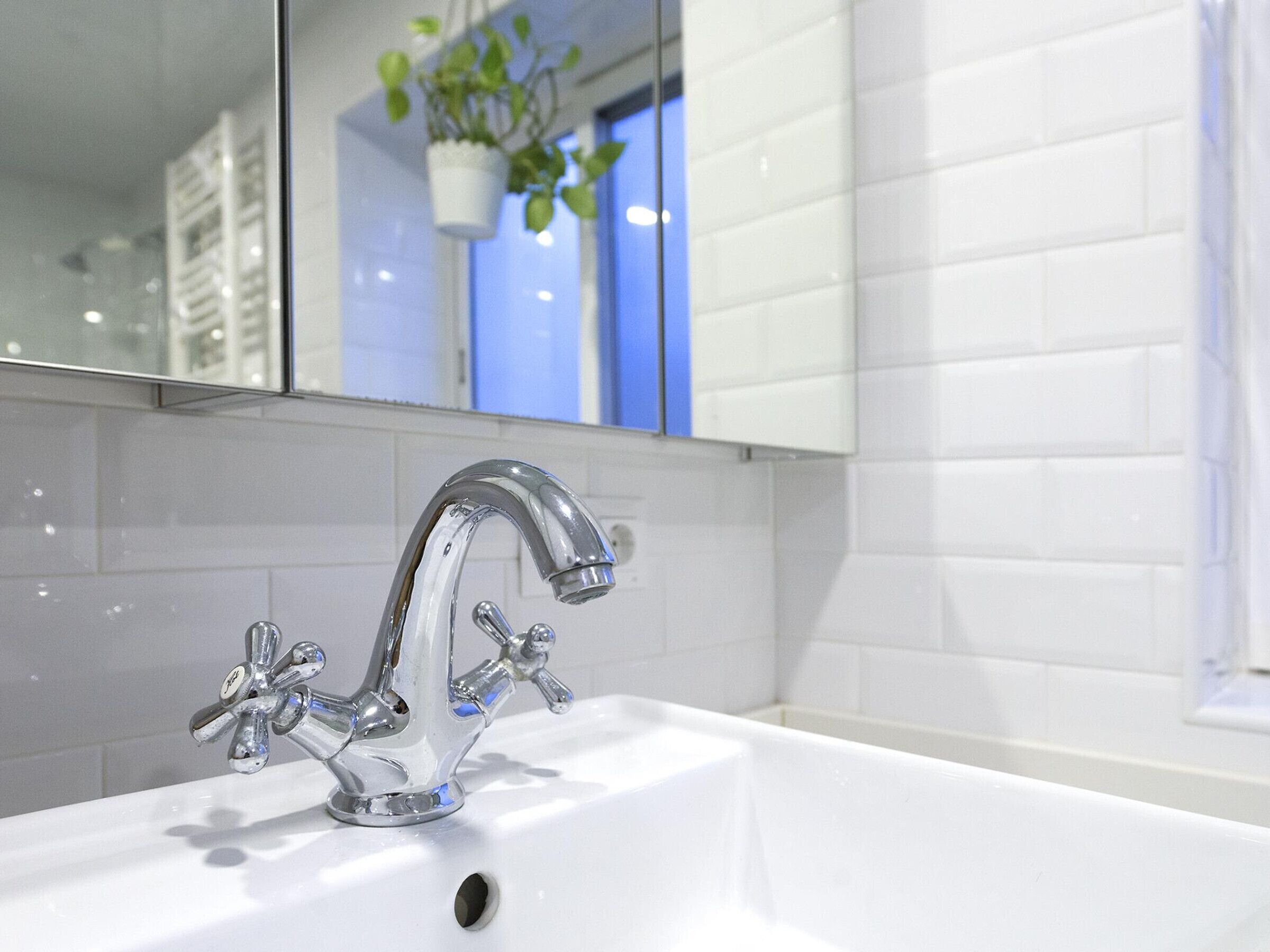Een moderne badkamer met een chromen kraan, witte tegels en een spiegel die een plant en een raam weerspiegelt.