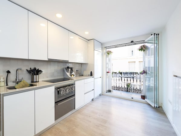 Una cocina moderna con armarios blancos, horno integrado y ventana con vistas a un balcón amueblado.