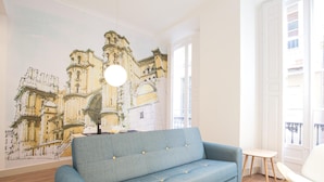 A living room with a blue sofa, a white coffee table, and a large mural of a historic building. - Del Parque Flats Santos Catedral - Cozy Apt in Málaga's Historic Center, Walk to Attractions (Málaga)